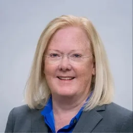 Headshot of woman with blonde hair wearing a gray jacket in front of a solid light gray background