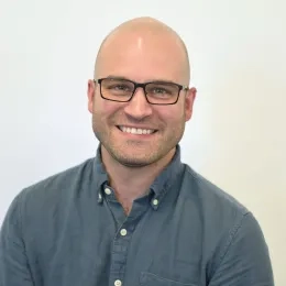 Headshot of man against a solid white background blue button-down shirt and glasses.