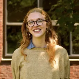 Headshot of Woman with long brown hair wearing glasses and a tan sweater in front of a brick building
