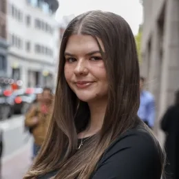 Headshot of woman with long brown hair wearing a black shirt in front of a city street in the background