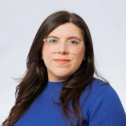 Headshot of woman with long black hair wearing glasses and a blue shirt against a solid gray background