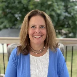 Headshot of woman with long brown hair wearing a white shirt, a blue sweater, and a necklace sitting on a balcony overlooking a lawn.