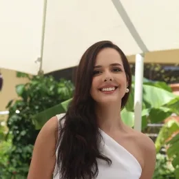 Headshot of woman with long black hair wearing a white dress with green plants in the background.