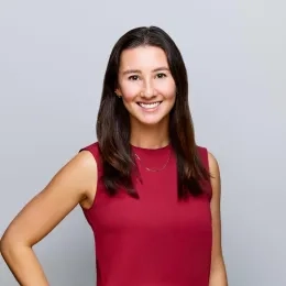 Headshot of woman with long dark hair wearing a red shirt against a gray background.