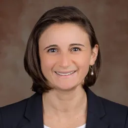 Headshot of woman wearing white shirt and black jacket against a brown backgroud
