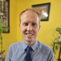 Headshot of Christopher Bodden wearing a blue button up shirt and blue tye in front of a yellow wall with paintings hung in the background.