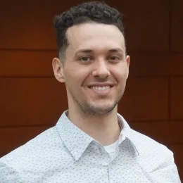 Man with short dark hair wearing a white patterned collared shirt in front of a red background