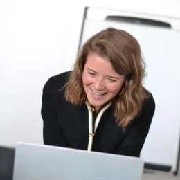Photo of Weyland with shoulder length brown hair standing in front of a Mac laptop wearing a black top