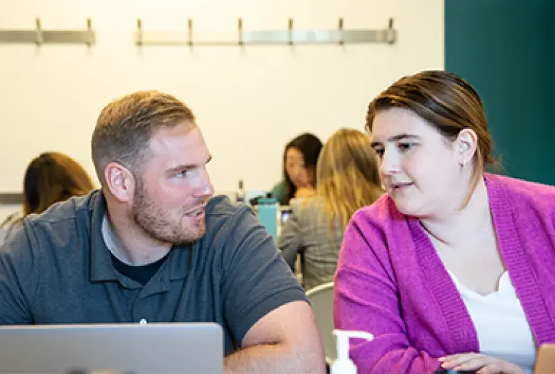 a woman leans over to look at a laptop while a man talks to her about something