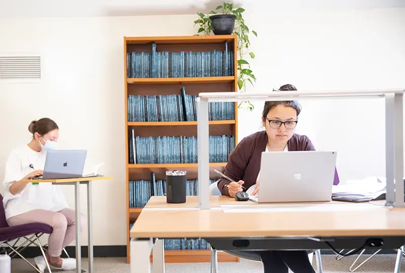 woman sits at laptop in front of a shelf of books while another works on a laptop at a table next to her