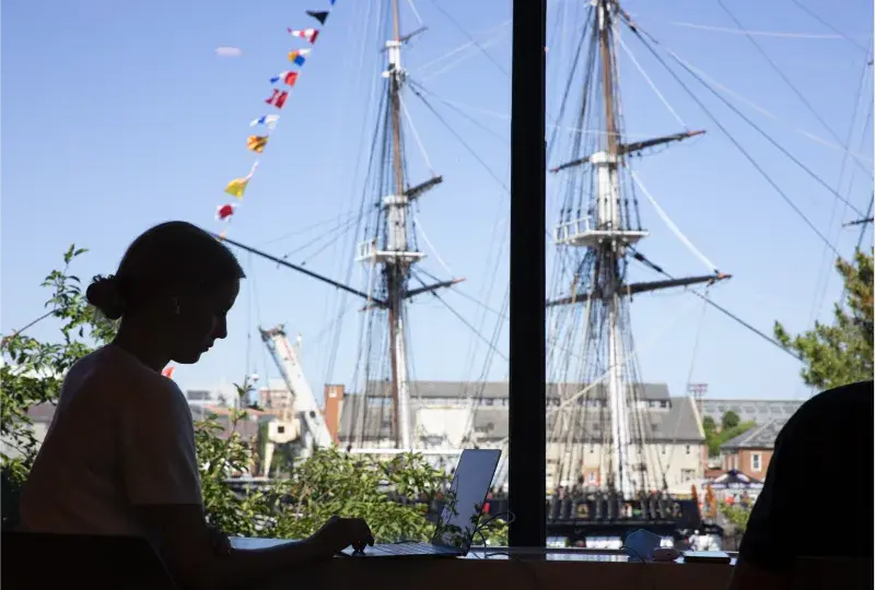 A person working on a laptop outside with a colonial ship in the back round
