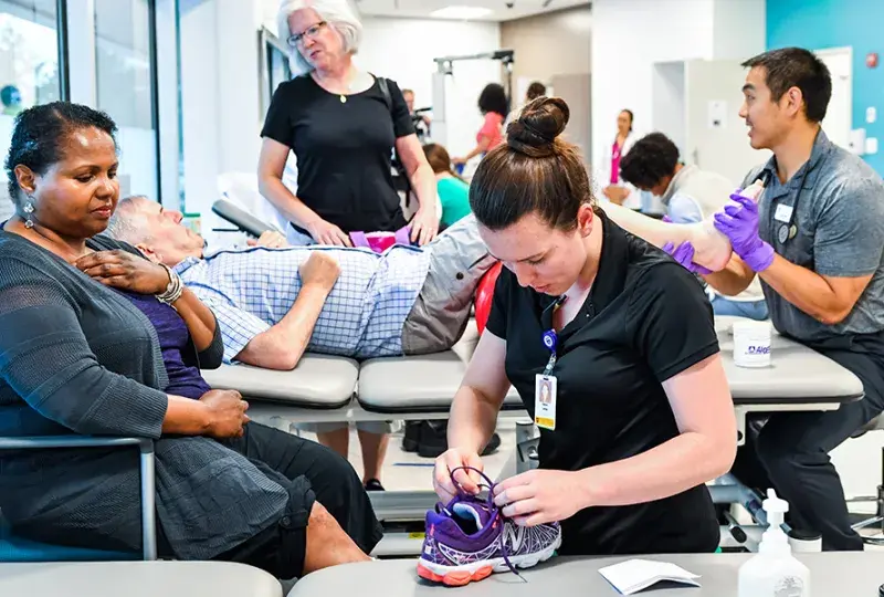 PT students work with patients, one ties a shoe the other rubs a foot