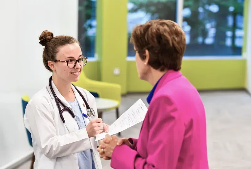 woman with glasses in a white lab coat speaks to a woman in pink
