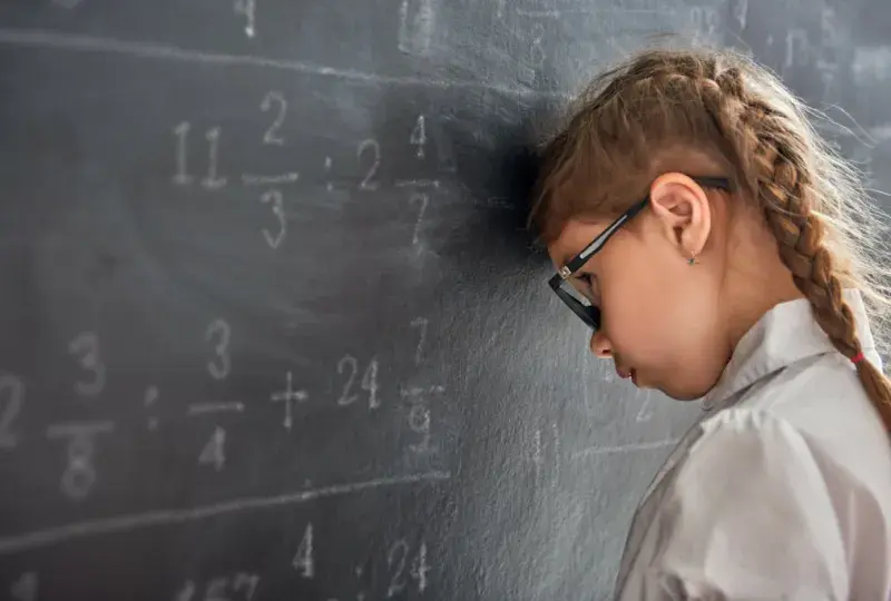 little girl with braids rests her head sadly against a chalkboard with math on it