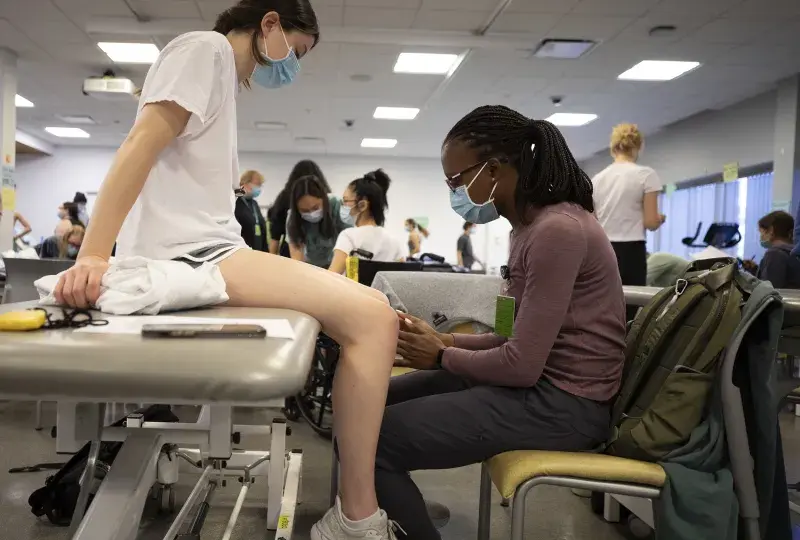 one student sits on an exam table, another touches her knee