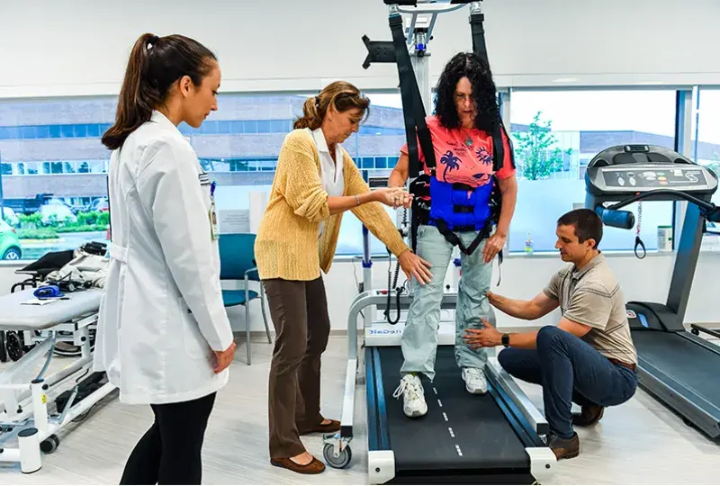 a woman in a harness walks on a treadmill helped by staff members