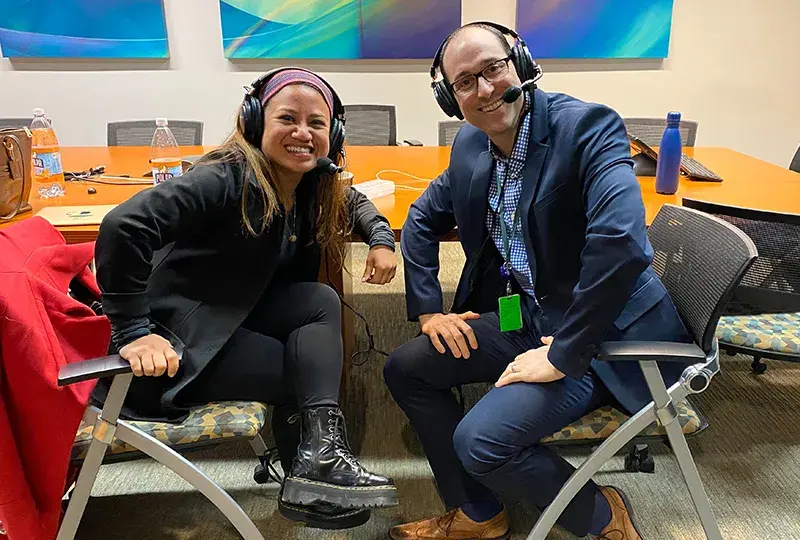 janice and peter sit in office chairs turned away from a table and smile at the camera wearing headphones