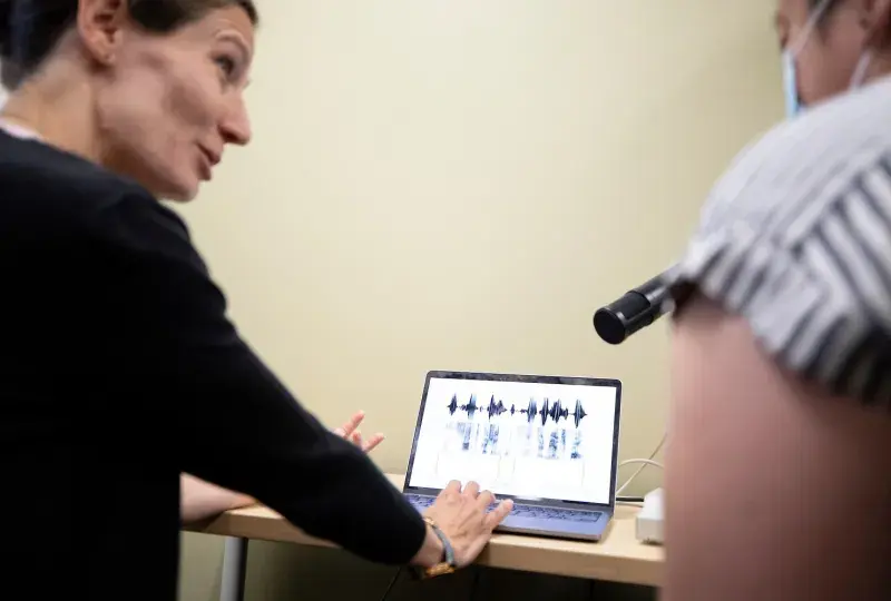 woman gestures to a laptop with soundwaves on it and a microphone in front