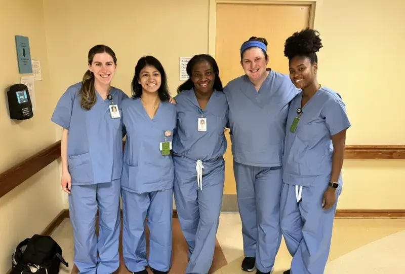 Five women wearing medical scrubs stand in a hallway posing for the camera