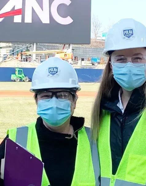 two women stand with ball field behind them wearing hard hats