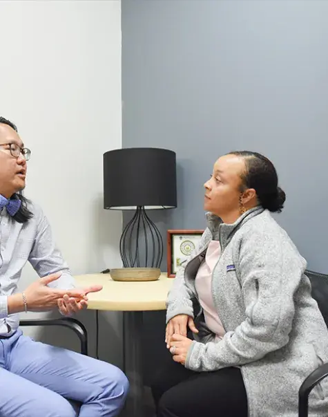 man of Asian descent sits and speaks with a woman with curly hair and brown skin