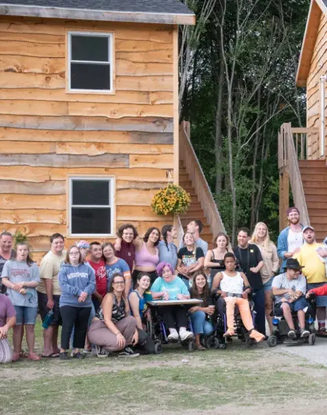 a large group of people stand and sit in wheelchairs in front of two wooden cabins
