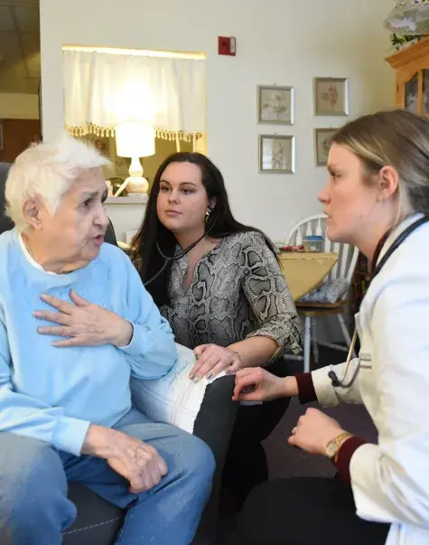 in a living room, an older woman with white hair puts a hand to her chest as two younger females crouch down to speak to her