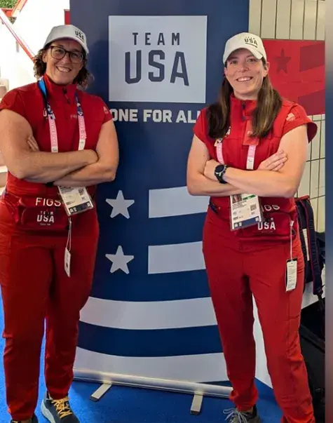 two women in red outfits pose in front of a team USA sign