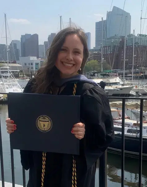 A woman wearing a graduation gown holds a diploma in front of a background of sailboats