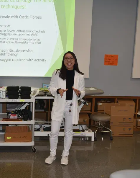 A woman in a white sweater and pants stands in front of a screen in a classroom with medical items behind her
