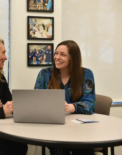 Two women sit at a desk talking with a computer between them
