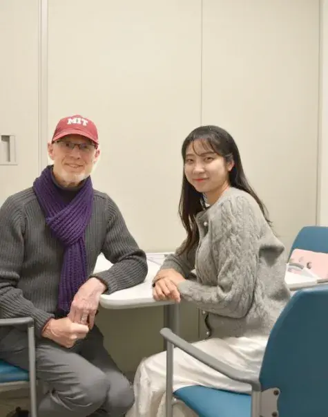 A man in a red baseball cap sits at a table facing the camera while a woman sits next to him facing the camera