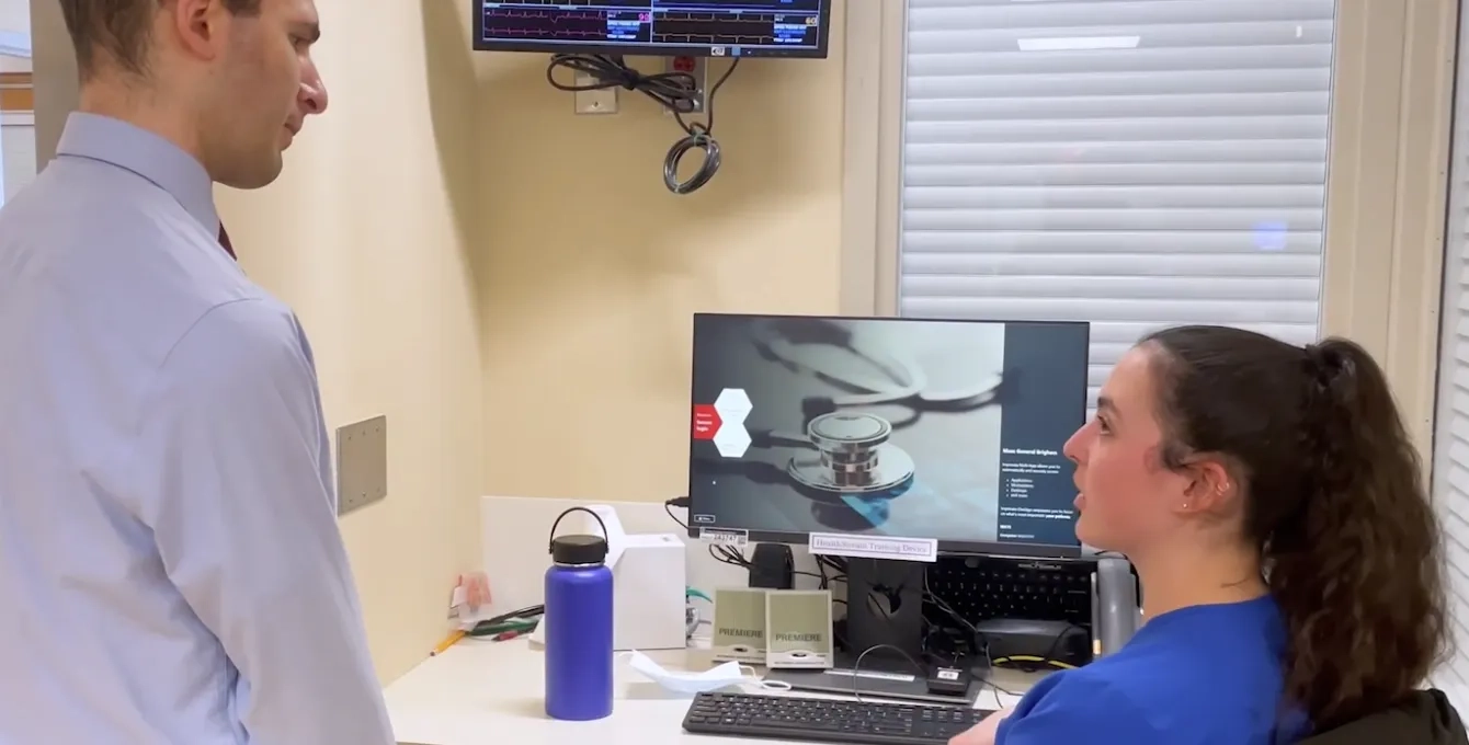 man in dress shirt speaks to a woman in scrubs at a computer