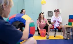 child in red sox jersey balancing on a stool smiles as a person in the foreground tosses a yellow ball to him